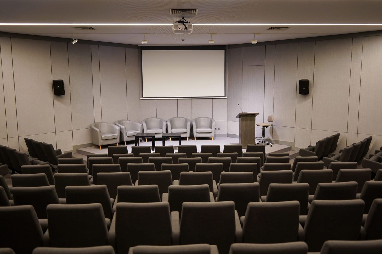 Leerer Konferenzsaal mit Stuhlreihen, Leinwand, Rednerpult und Sesseln auf der Bühne
