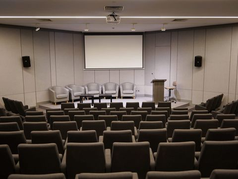 Leerer Konferenzsaal mit Stuhlreihen, Leinwand, Rednerpult und Sesseln auf der Bühne