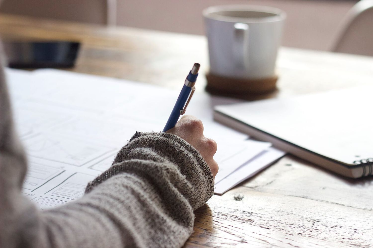 Hand skizziert mit Druckbleistift Website-Wireframes auf Papier, daneben Kaffeetasse und Notizbuch